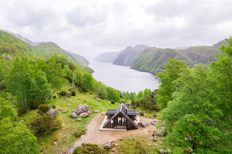 En lang fjord omgitt av fjell, grønne store trær og en dagsturhytte som er plassert helt ytterst på kanten av fjellet slik at utsikten til fjorden åpenbarer seg bak.