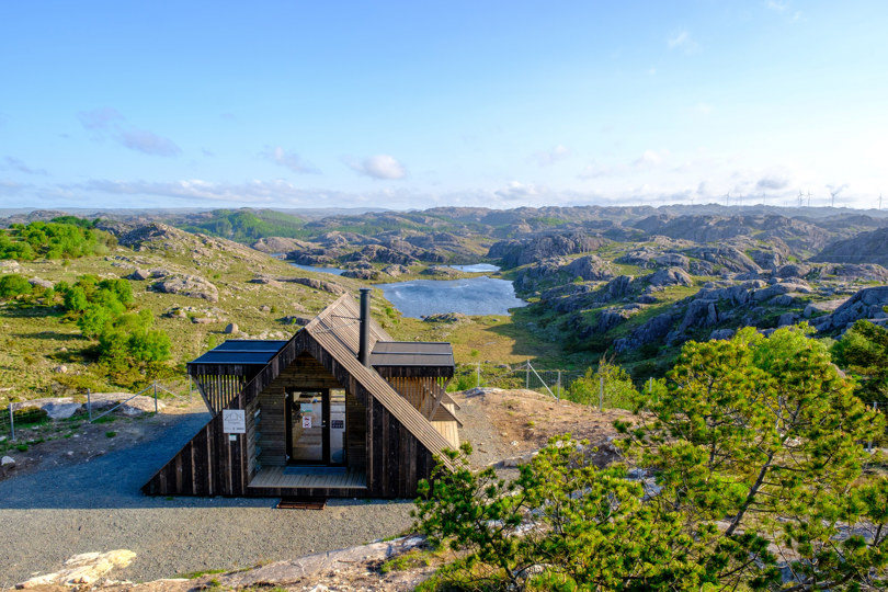 En brun hytte som ligger plassert på et gruslagt område som er planert. Terrenget rundt består av trær, litt kupert terreng med berg. Havet skimtes i det fjerne.