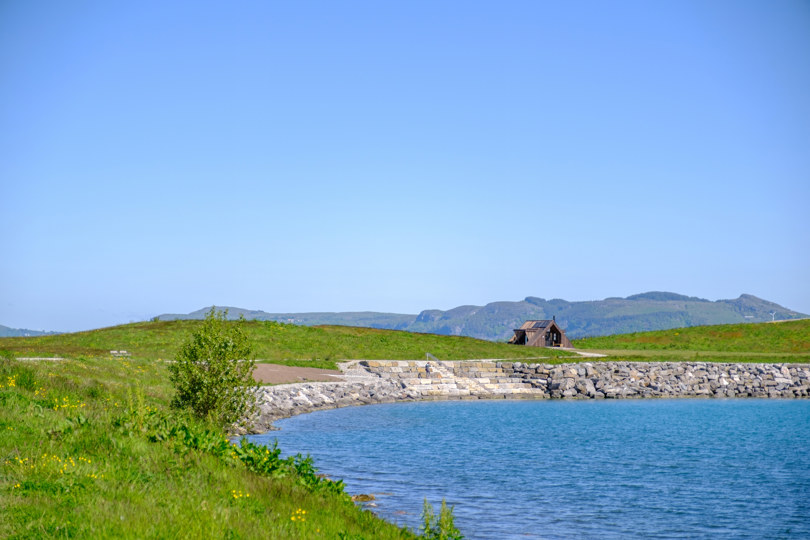 Dagsturhytte som ligger ytterst på et nes. Langs neset er det en steinmur og og blått vann. I fronten av bildet vises grønt gress. Fjell vises i horisonten og himmelen er knallblå.