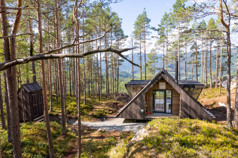 En dagsturhytte som ligger midt inne i en skog omgitt av høye furtrær. I bakgrunnen kan du se utsikten til Suldalsosen.