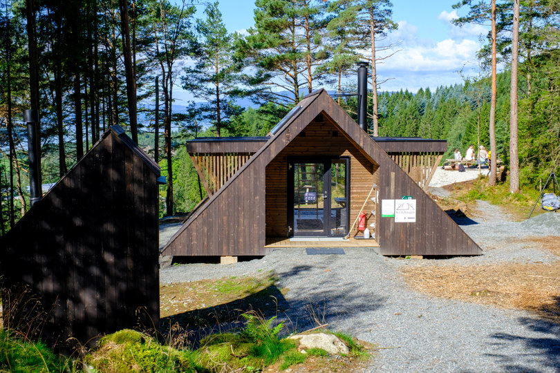 En dagsturhytte som ligger plassert på et gruslagt område inne i en skog. Stien er gruslagt inn til hytta. I front av bildet vises et brunt lite tilbygg som muligens er toalettet som er tilknyttet hytta.