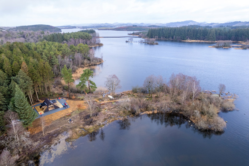 Oversiktsbilde som viser et vann, et nes, skog og en dagsturhytte som ligger plasser rett ved skogen og ytterst mot vannet.