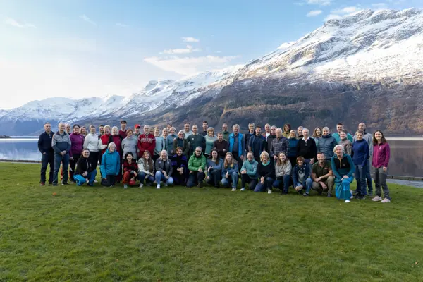 En gruppe mennesker står på en gressplen med fjord og snøkledd fjell bak seg.