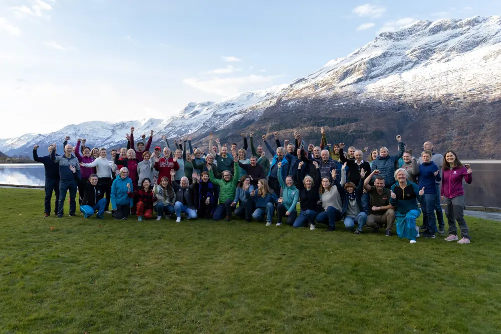 En stor gruppe mennesker smiler foran snøkledd fjell og fjord i bakgrunnen.