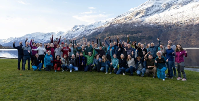 En stor gruppe mennesker smiler foran snøkledd fjell og fjord i bakgrunnen.