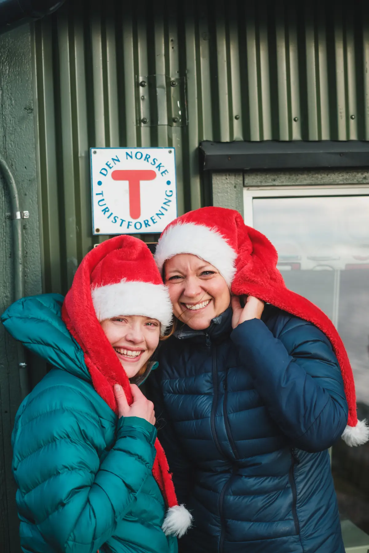 To damer med julenisseluer foran dnt-hytte med T-skilt.