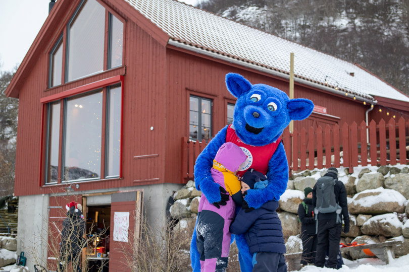 En stor rød løe i bakgrunnen av bildet med snødekt landskap. En utkledd maskot i blå revekostyme klemmer på to barn.