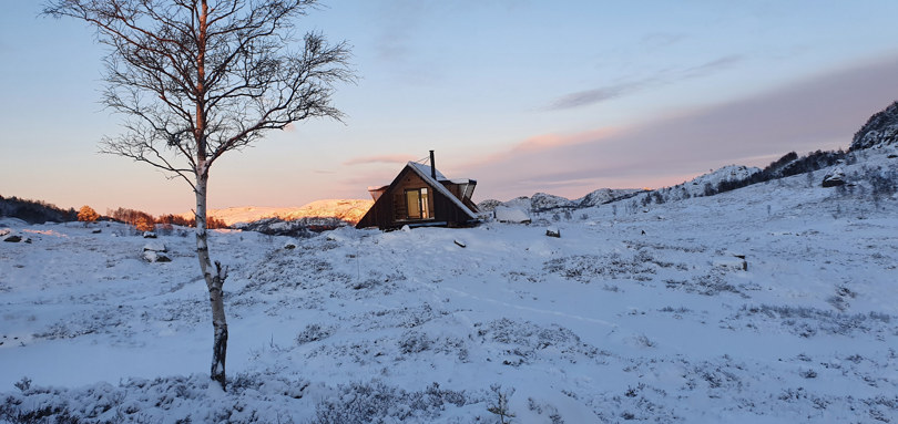 Snødekt landskap med en trekantet dagsturhytte i det fjerne. Solnedgang. Et tre med snø vises i fronten av bildet.