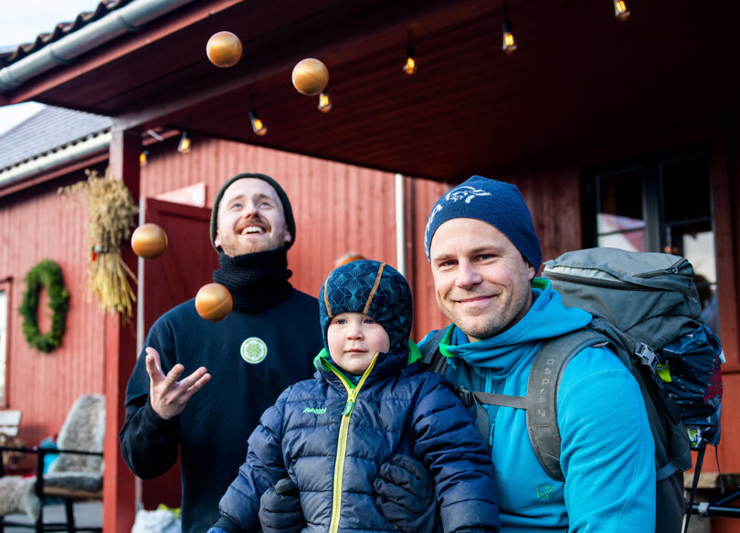 En sjonglør sjongelerer med tre baller i bakgrunnen mens en far og en sønn sitter på huk og smiler til kamera i front.