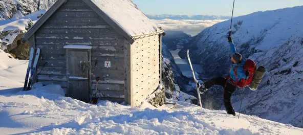 En mann i blå boblejakke jubler ved hytten Kiellandbu i vinterlandskap med fjord bak seg.
