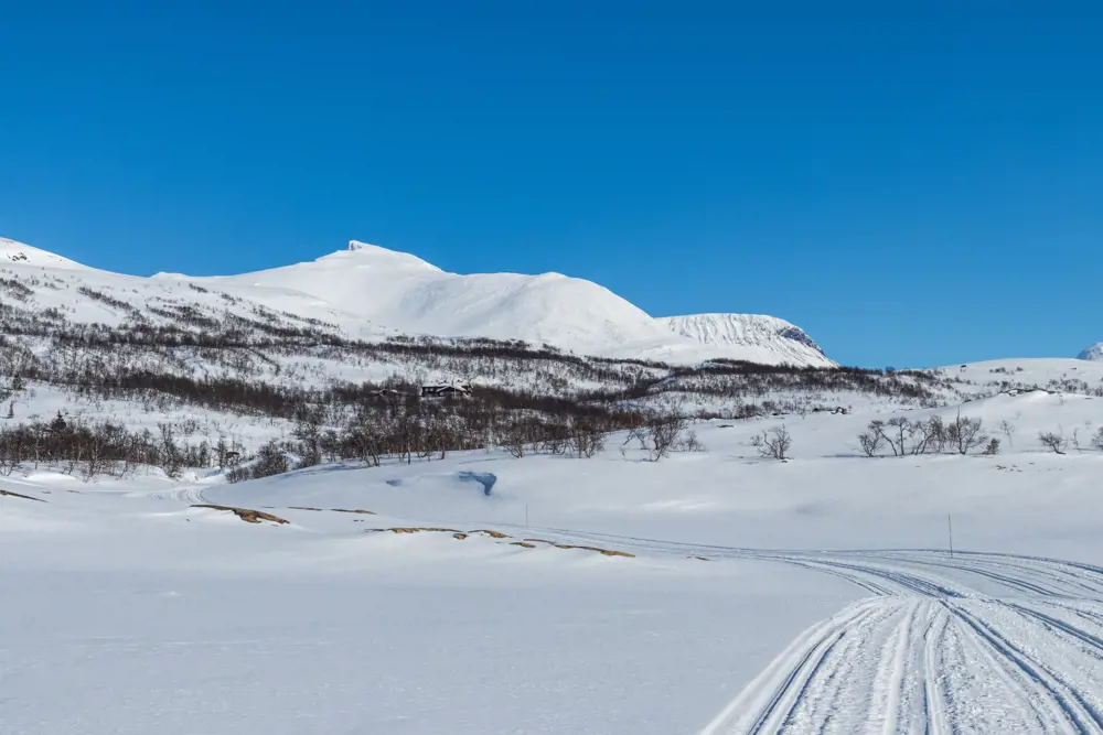 Store fjell, helt snødekt, ruver opp under blå himmel. Fotografert fra et islagt vann med masse snø, og snøscooterspor leder opp fra vannet og opp i skogen til en hytte ved foten av fjellene.