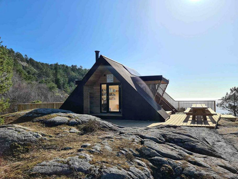 Et nærbilde av en trekantet dagsturhytte som ligger plassert på et berg. Treterrasse utenfor hytta med en stor trebenk hvor man kan sette seg ned og nyte omgivelsene.