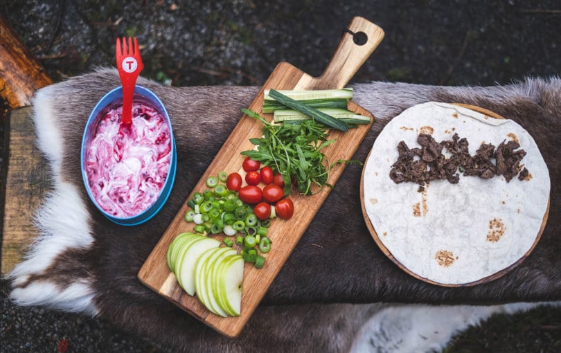 Turmat presentert med en lefse med viltkjøtt, en fjøl med oppskåret eple, tomat, vårløk, agurk og salat, samt en boks med tyttebærrømme.