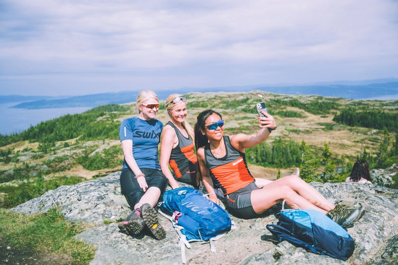 Tre kvinner sitter på bakken på en steinete topp, kledd i lette turklær, mens den forreste av dem tar en selfie av alle tre.