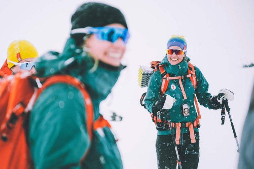 To personer kledd i den samme mørke turkise vingerjakken, og med den samme oransje tursekken, med snøbriller, står og smiler til kamera, mens lett snø faller rundt dem. Personen til høyre holder i skistaver.