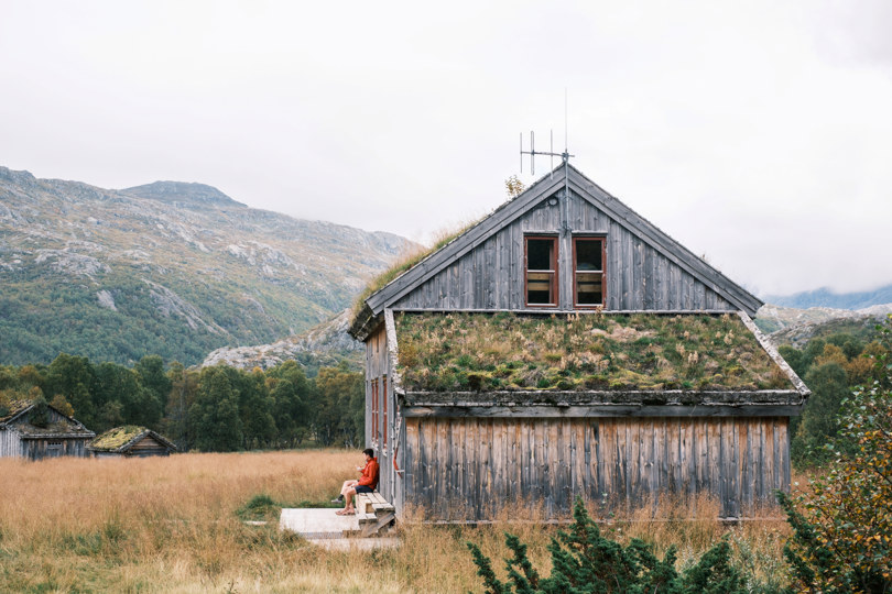 Grå, eldre hytte som ligger i et åpent landskap med siv, og høye fjell. En person i rød genser sitter på en benk utenfor hytta.