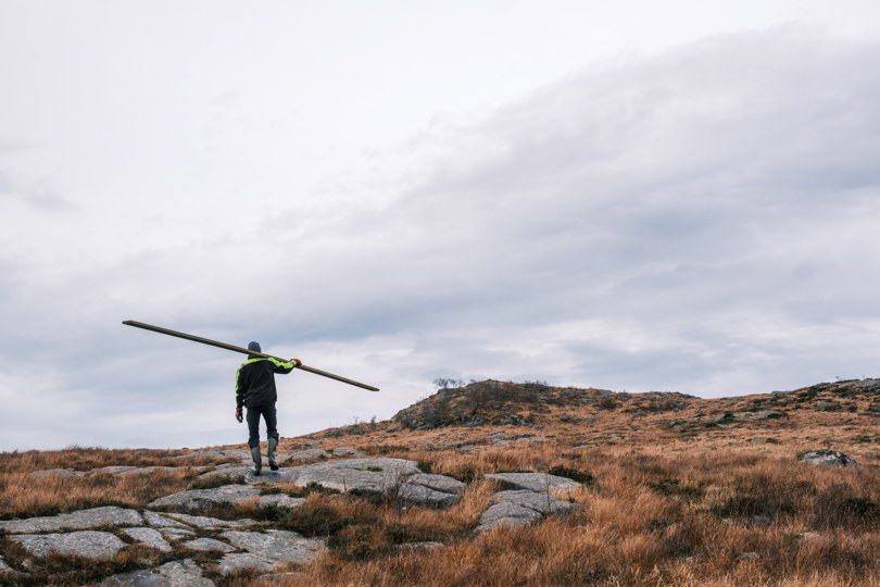 Mann bærer planke på skulderen i brunt fjellandskap.