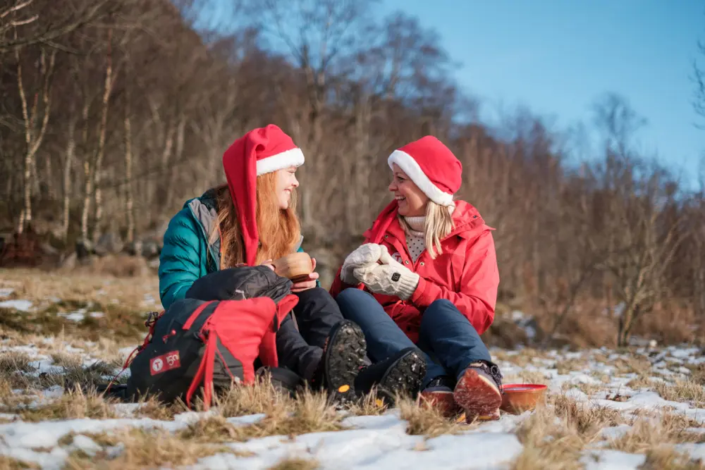 To damer sitter i snødekt gress med nisseluer. De ler og koser seg mens de drikker kaffe og spiser pepperkaker.