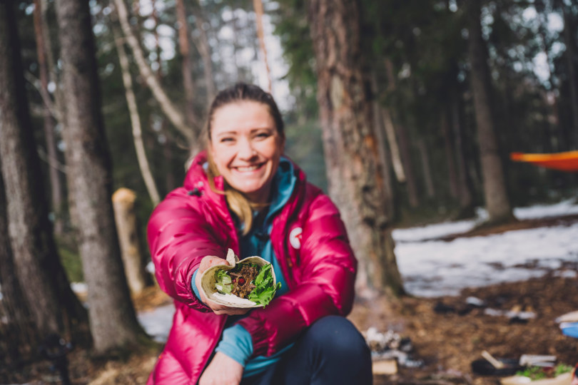 En kvinne i en skinnende lilla boblejakke sitter på huk og tilbyr kameraet noen form for mat, met en skog synlig bak henne