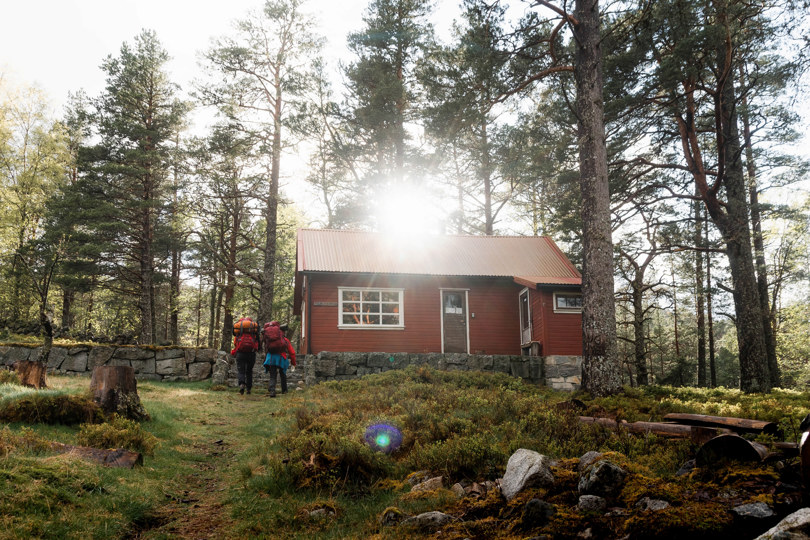 En rød hytte inni en skog. To personer med sekk går opp bakken mot hytta.