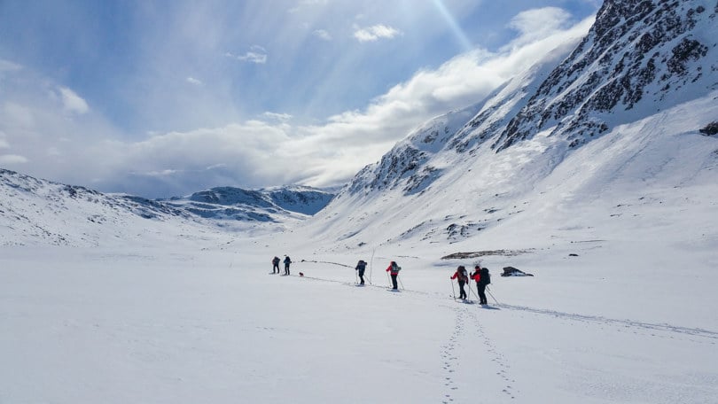 Seks personer, delt opp i tre grupper, går på ski langs det samme skisporet, i et stort og øde snødekket landskap, grenset av et fjell på høyre side.
