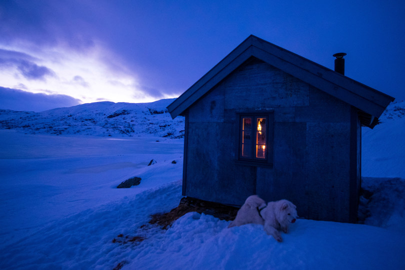 Grå hytte i kveldslys med snødekt landskap.