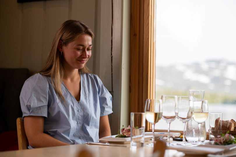 Kvinne med lyst hår og lyseblå bluse sitter ved et bord og nyter en bedre middag.