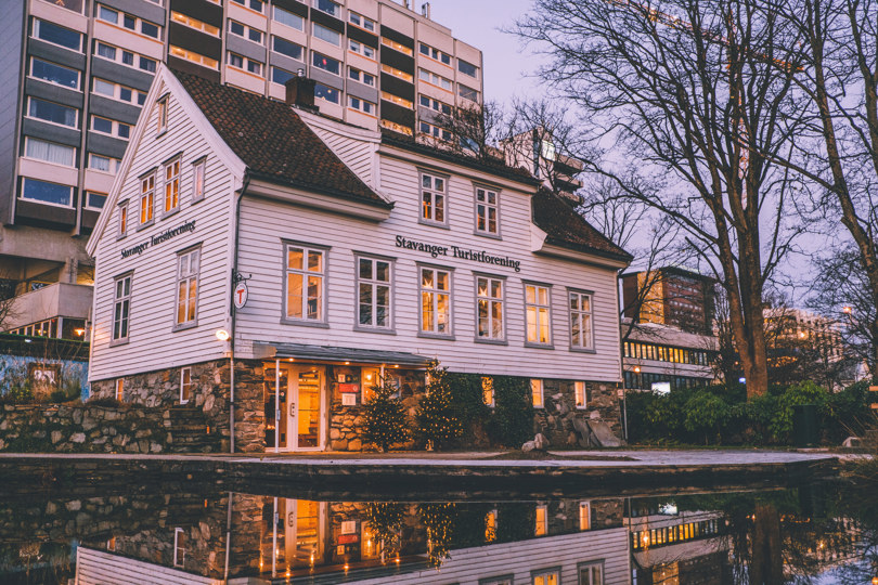 Hvitt hus med Stavanger Turistforening sitt navn på veggen som speiler seg i en dam. Julebelysning og hvitt på bakken.