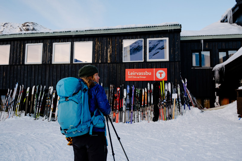 Dame som står foran en hytte hvor det er lent mange ski inntil veggen