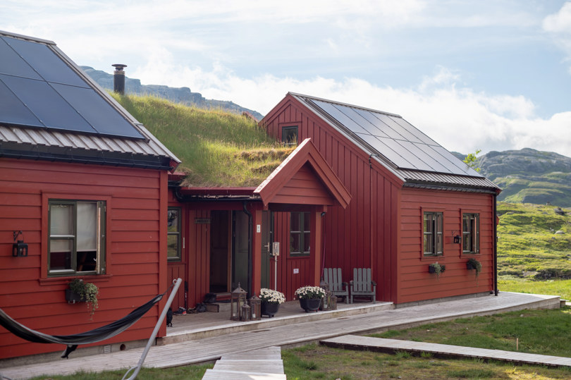 Rød hytte med solceller på taket, blomster utenfor og en hengehøye som henger til venstre i bildet.