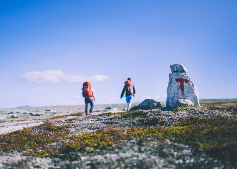 Stein med rødmalt T på høyfjellet. I bakgrunnen skimtes to turgåere.