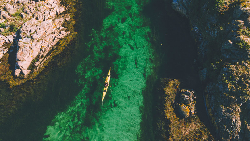 Fugleperspektiv av en person som padler i gul kajakk i grønt vann mellom to skjær.