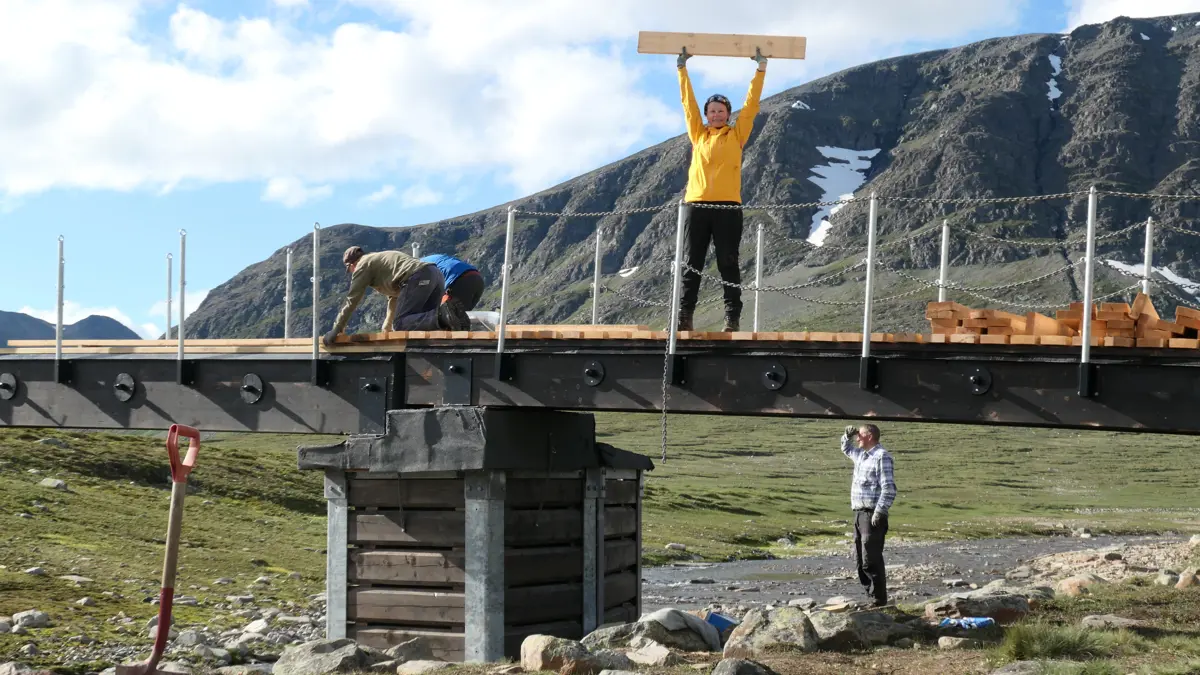 En bru er i ferd med å bli bygget. Fire personer er synlige, en som står på brua og holder en planke over hodet, to som er på alle fire og jobber på brua, og en som står på bredden av elva under brua.