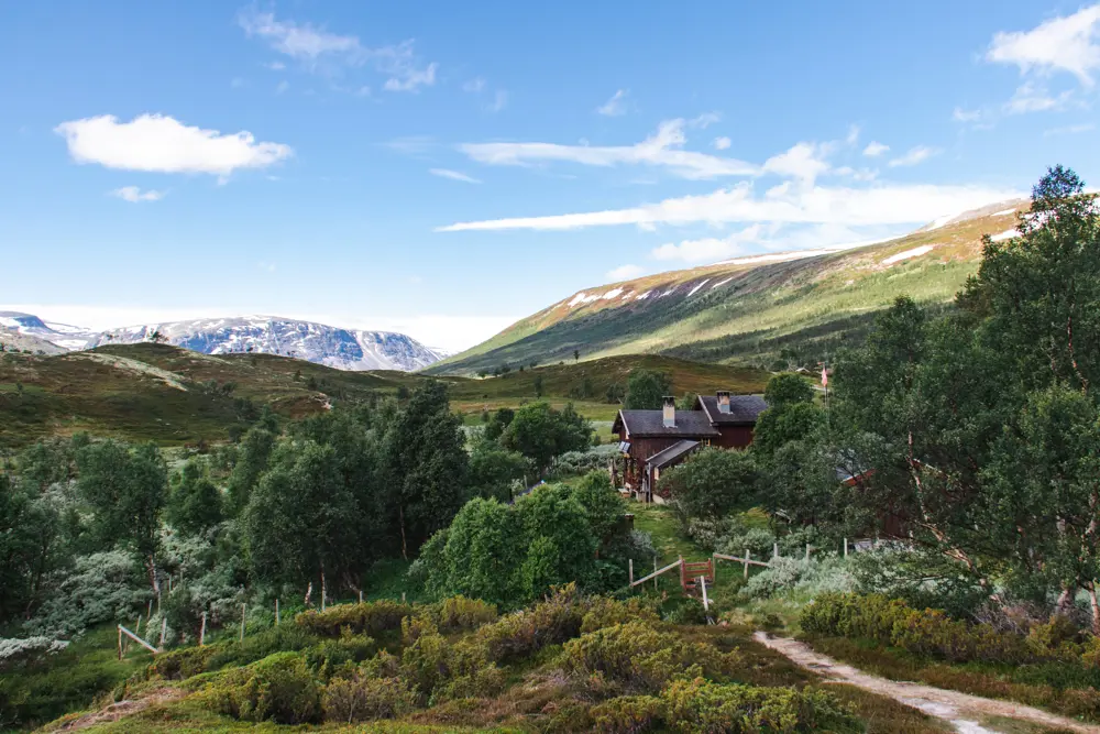 Bildet viser en åpen fjelldal med mye lyngterreng og noe buskvekst. En sti fører ned til en liten hytte, og i bakgrunnen ruver store fjell.