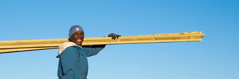 Smilende mann med grønn jakke og blå lue  holder noen planker mens det er blå himmel i bakgrunnen