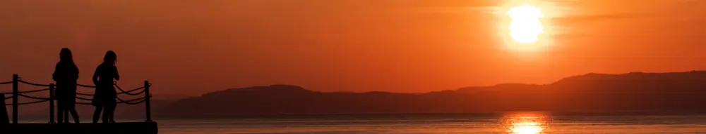 Bildet viser silhuetten av to figurer og kaia de står på i venstre bunn-hjørne, mens resten av bildet viser en solnedgang over fjorden, med landmassen på andre siden av fjorden synlig i det fjerne.