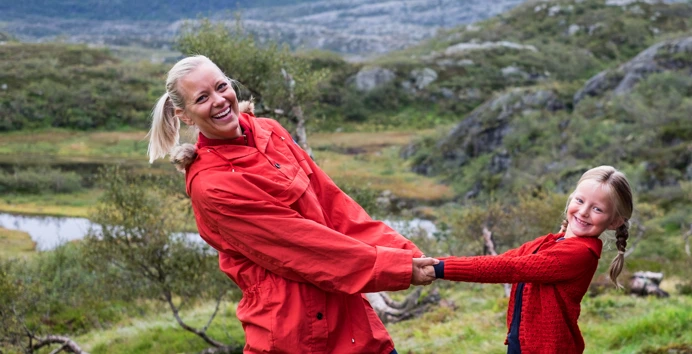 Mor og datter i rødfargede klær holder hender, lener seg tilbake og smiler mot kamera. Landskapet er grønt og på fjellet.