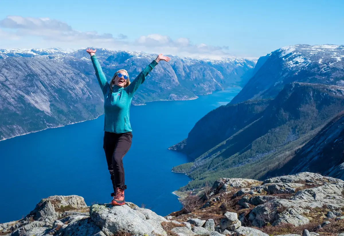 Dame står på toppen av et gjell med armene i luften. Bak er det fjord og fjell