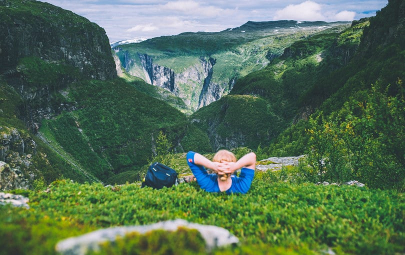 Mann i blå genser ligger i lyngen med hendene bak hodet og skuer ut over en grønn og frodig dal med bratte fjellsider..