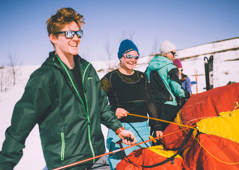 To personer setter opp et rødt og gult telt solskinn på vinterfjellet.