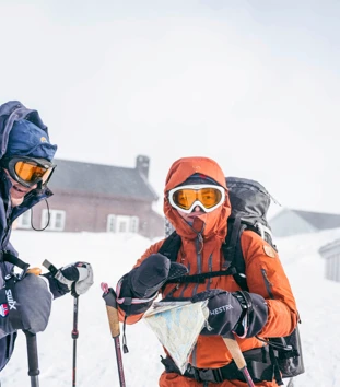 To skiløpere i vinterfjellet, i sterk vind, med hette, briller og kart.