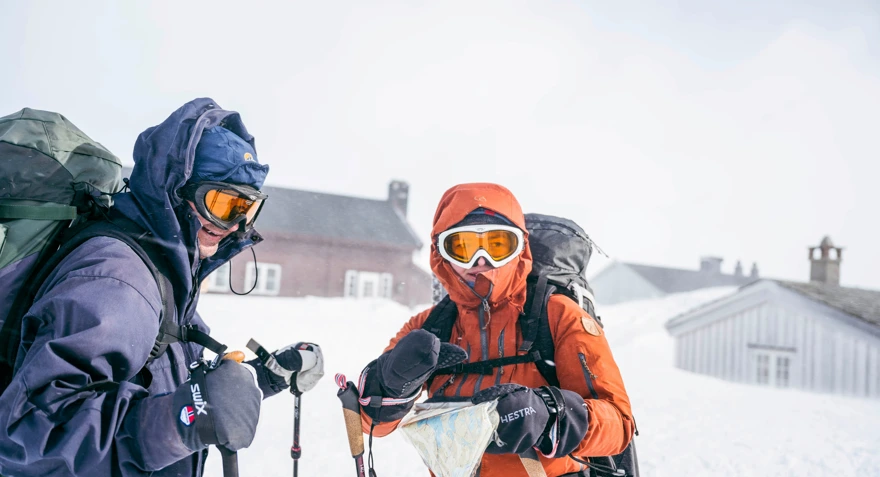 To skiløpere i vinterfjellet, i sterk vind, med hette, briller og kart.