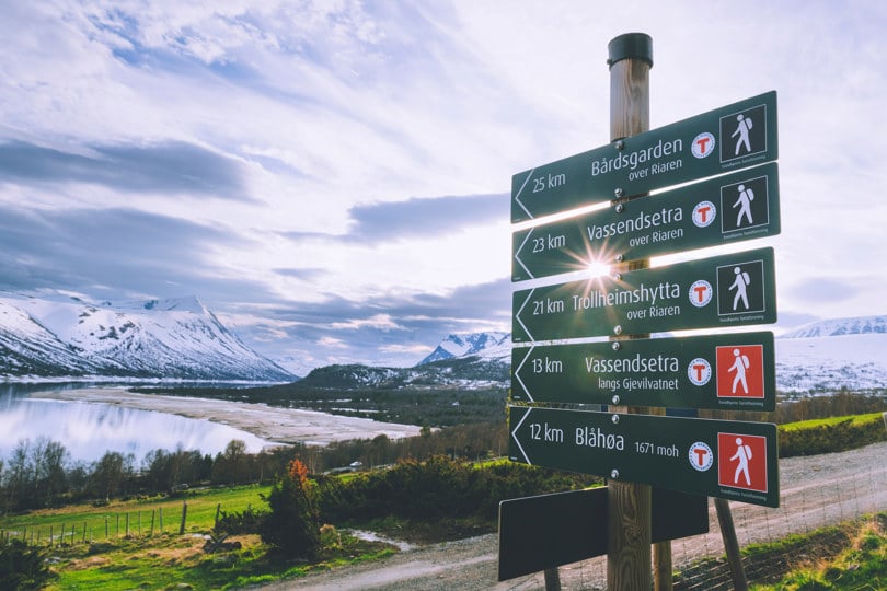 Bildet viser en skilt-stav, der fem forskjellige skilt peker veien og avstanden til andre lokasjoner. Fra toppen: Bårdsgaden, Vassendsetra (over Riaren), Trollheimshytta, Vassendsetra (langs Gjevilvatnet), og Blåhøa. Delvis snødekket fjell i bakgrunnen, og mellom oss og der er det en innsjø. Solen skinner mellom to av skiltene. 