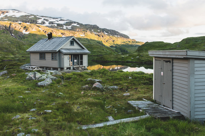 Grå plankekledd hytte og uthus ved siden av et lite fjellvann. 