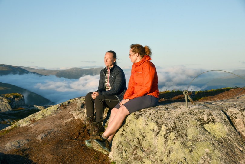 To kvinner i svart og rød jakke sitter på en fjelltopp. 