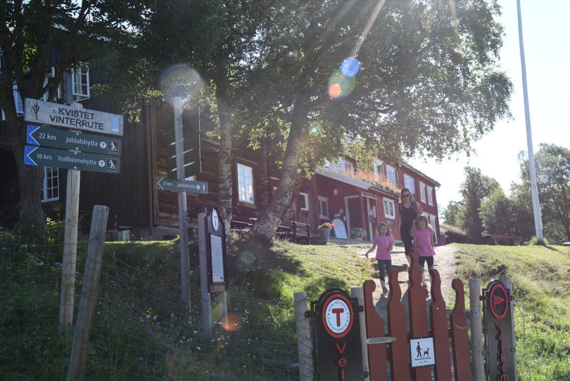 Tre personer går nedover en liten bakke mot ei grind foran ei stor hytte.