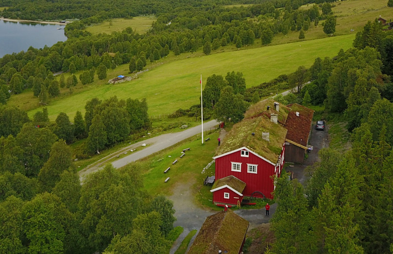 Fugleperspektiv av rød hytte med torvtak. Rundt hytta er det grønn skog og grønne enger.