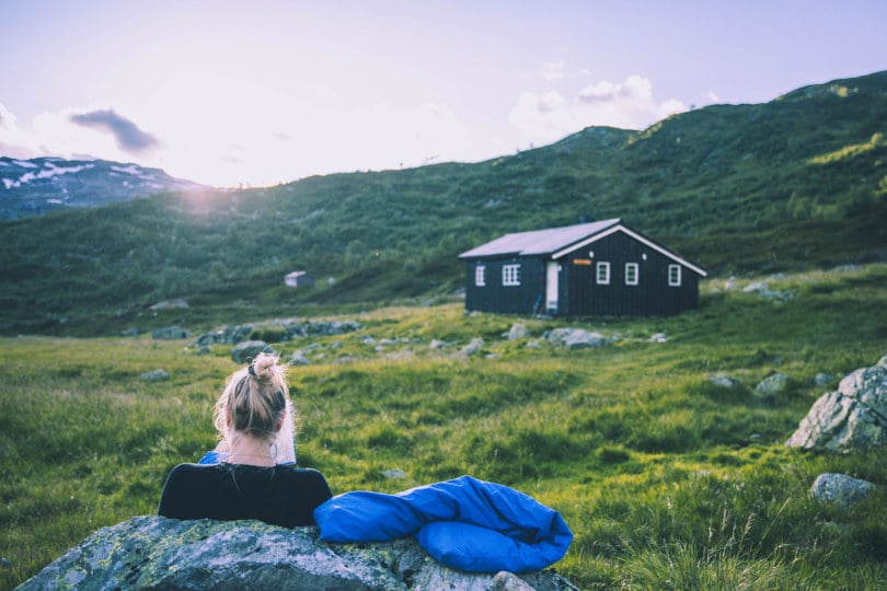 Kvinne sitter inntil en stein med ryggen til og leser ei bok i nærheten av ei svart hytte i en grønnkledd fjellside.