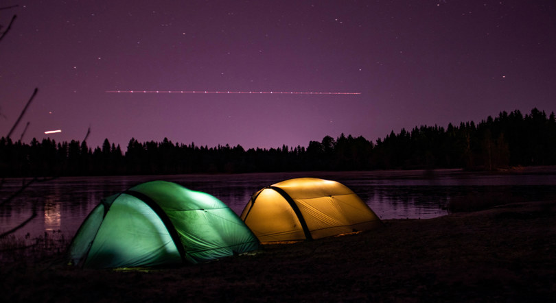 To telt lyser opp i forskjellige farger ved et tjern i skogen. Det er mørkt, og på himmelen ser man stjerner og lys etter et fly.