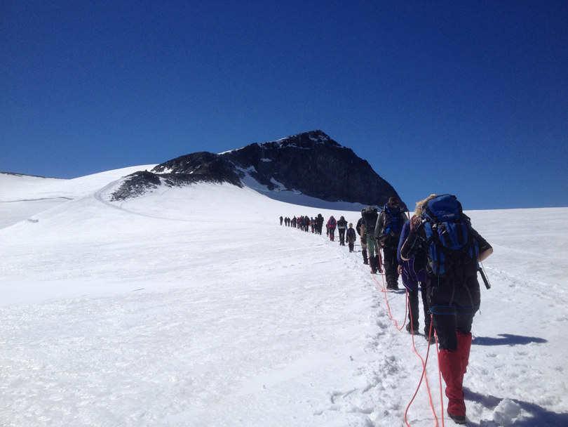 En lang rekke med mennesker går over en bre på vei mot en fjelltopp.